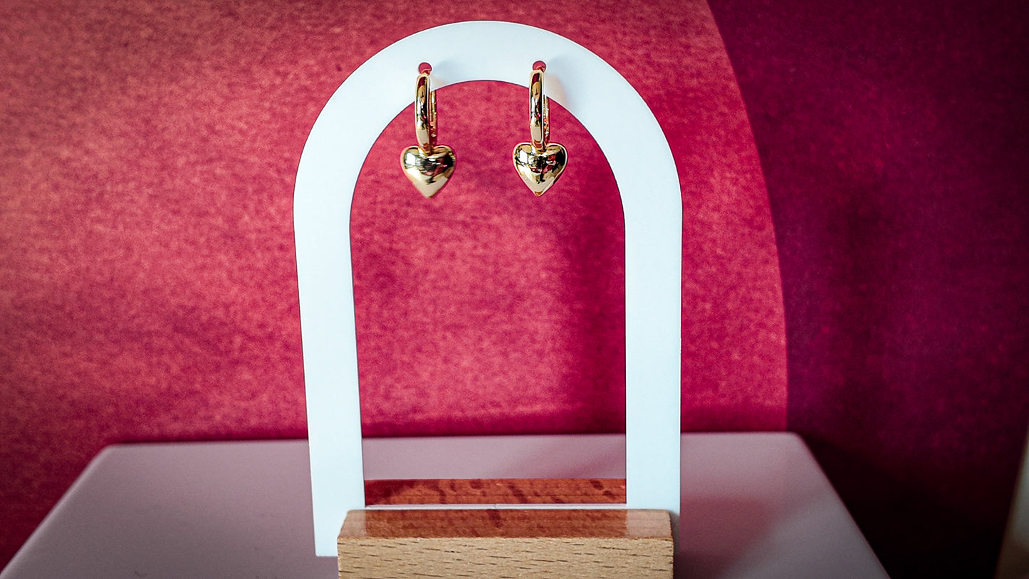 Gold heart-shaped earrings on a white arch against a pink background