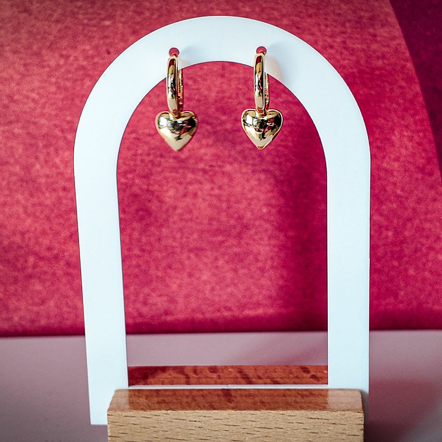 Gold heart-shaped earrings on a white arch against a pink background