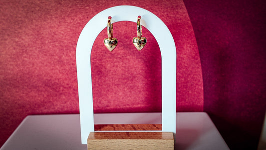Gold heart-shaped earrings on a white arch against a pink background
