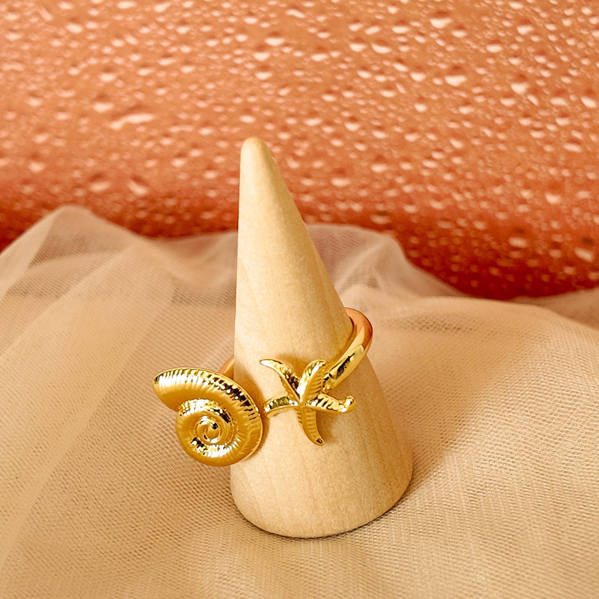 Gold ring with decorative elements on a beige cone-shaped stand against a textured pink background.