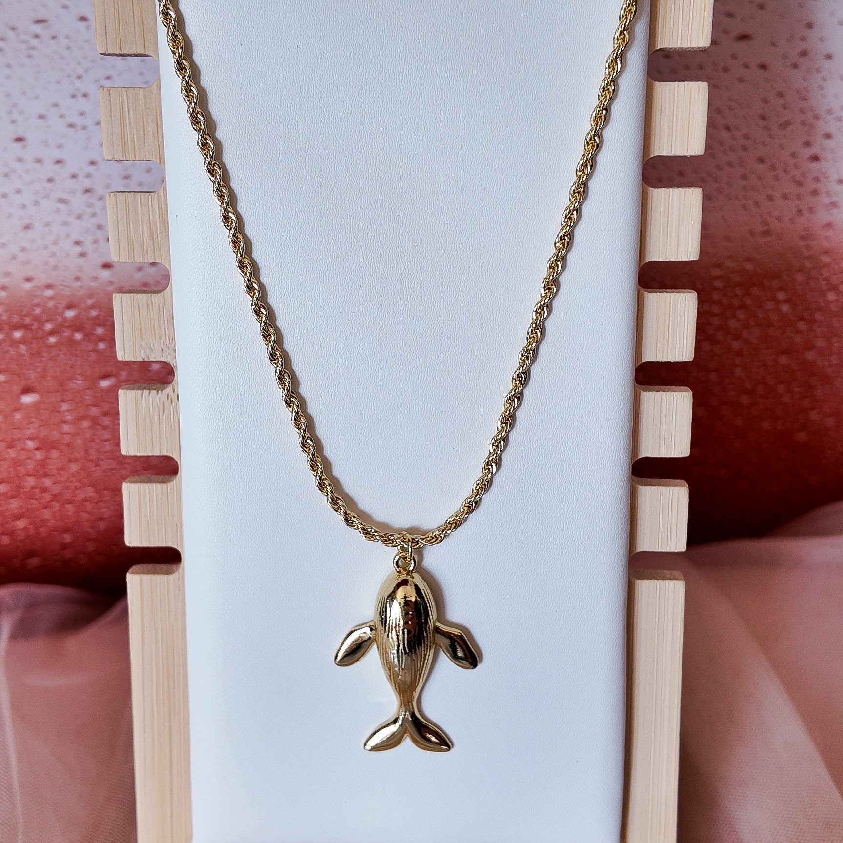 Gold necklace with a dolphin pendant on a white card against a pink and wooden background
